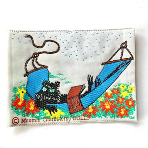Puede incluir: Parche bordado que representa un personaje de dibujos animados negro relajándose en una hamaca azul, leyendo un libro. La escena incluye flores coloridas y el texto "Moomin Characters/BULLS". El parche tiene un fondo blanco.