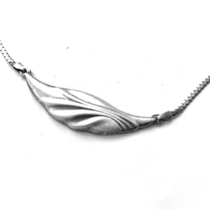 May include: A silver necklace with a leaf-shaped pendant. The pendant has a curved, elongated design with detailed veining. The necklace chain is a delicate, box-style link. The jewellery is set against a white background.