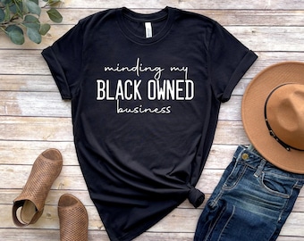 May include: Black t-shirt with white text that reads "minding my BLACK OWNED business". The shirt is laying on a wood surface with a brown hat, blue jeans, and brown sandals.