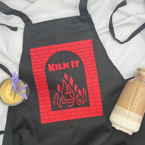 May include: Black apron with a red brick design and the text "Kiln It" in a white archway. A flame design is inside the archway.