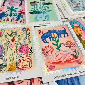 May include: A collection of rectangular tarot cards featuring colorful hand-drawn illustrations with holographic silver borders. The cards display various figures, animals, and symbolic imagery with text labels including Free Spirit, Flourishing, and Dreamer and the Dream.