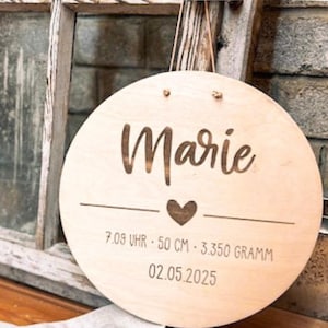 May include: A round wooden sign with the name "Marie" engraved in a flowing script. Below the name is a heart symbol and the measurements: 7:09, 50 cm, 3.350 grams, and the date 02.05.2025. The sign is light-colored with a natural wood grain.