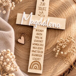 Può includere: Un ornamento a forma di croce in legno chiaro con la scritta "ZUR TAUFE" e "Magdalena" in lettere bianche 3D. La croce presenta l'illustrazione di una colomba, una poesia in tedesco e un arcobaleno sul fondo, appesa a un sottile spago.