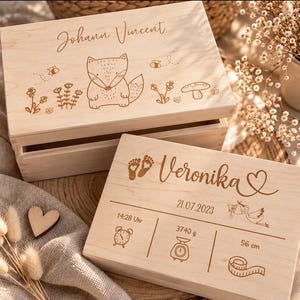 Può includere: Due scatole rettangolari in legno chiaro. La scatola superiore presenta una volpe incisa, fiori e il nome Johann Vincent. La scatola inferiore mostra il nome Veronika, impronte, un cuore, una cicogna e i dettagli di nascita: 21.07.2023, 14:28, 3740 g e 56 cm.