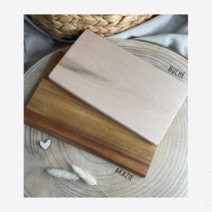 Pu&ograve; includere: Due taglieri rettangolari in legno impilati l'uno sull'altro. Il tagliere superiore &egrave; beige chiaro con il testo BUCHE stampato accanto. Il tagliere inferiore &egrave; in legno marrone pi&ugrave; scuro con il testo AKAZIE stampato accanto. &Egrave; visibile un piccolo cuore bianco.
