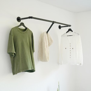 Könnte beinhalten: Eine wandmontierte Kleiderstange aus schwarzen Industrierohren mit einer horizontalen Stange und einem Stützarm. Die Stange trägt ein grünes T-Shirt auf einem schwarzen Bügel, einen cremefarbenen Strickschal und ein weißes Hemd auf einem schwarzen Bügel.