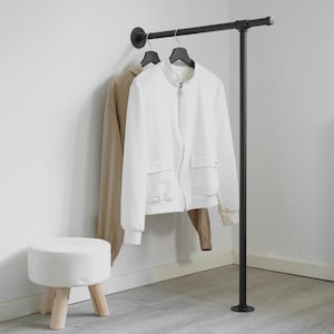 Könnte beinhalten: Eine weiße Jacke und ein beiger Kapuzenpullover hängen an einer schwarzen Metall-Kleiderstange. Ein kleiner, weißer, runder Hocker mit Holzbeinen steht vor der Stange. Die Stange steht vor einer weißen Wand.