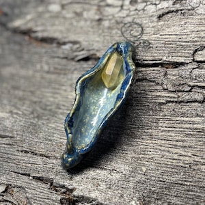 May include: A handcrafted ceramic pendant in the shape of a shell, with a blue and gold glaze. A yellow crystal is nestled inside. The pendant is displayed on a weathered wooden surface, showcasing its unique, organic form.