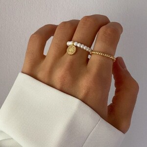 Könnte beinhalten: Zwei goldene Ringe an einer Hand. Ein Ring ist ein schlichtes Band aus kleinen goldenen Perlen. Der andere Ring ist eine Kette aus weißen Perlen mit einem goldenen Anhänger.