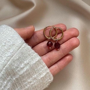 Pode incluir: Brincos de argola dourados com pingentes de pedras preciosas vermelhas.