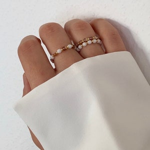 Könnte beinhalten: Zwei Perlenringe an einem Finger. Die Ringe bestehen aus kleinen, weißen und braunen Perlen. Die Perlen sind auf einem dünnen goldenen Draht aufgefädelt.