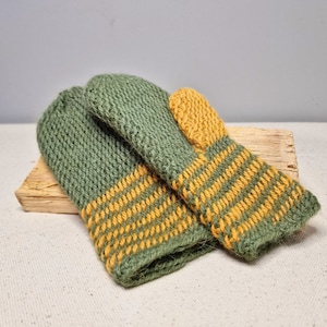 Könnte beinhalten: Ein Paar gestrickte Fäustlinge in Olivgrün und Gelb. Die Fäustlinge sind aus einem strukturierten Garn gefertigt, mit einem einfarbigen grünen Oberteil und einem gelb-grünen Streifenmuster auf der unteren Hälfte. Die Fäustlinge liegen auf einem Holzstück.