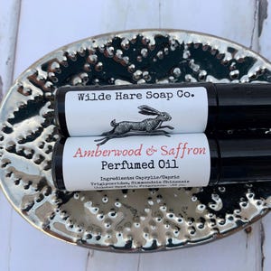 May include: Two black roll-on perfume bottles with white labels are stacked on a silver, textured dish. The top label reads "Wilde Hare Soap Co." with a rabbit illustration. The bottom label says "Amberwood & Saffron Perfumed Oil."