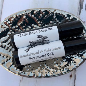 May include: Two small black roll-on bottles with white labels. The label on the top bottle reads "Wilde Hare Soap Co." and features a black and white illustration of a hare. The label on the bottom bottle reads "Sandalwood & Palo Santo Perfumed Oil" and includes the text "Ingredients: Caprylis/Capric"
