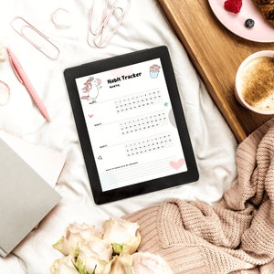 The digital habit tracker in the unicorn style of the monthly habit trackers for submissives - sitting on a bed next to coffee and a pink plate on a wooden tray, a knitted sweater, a gray notebook, and pink pen.