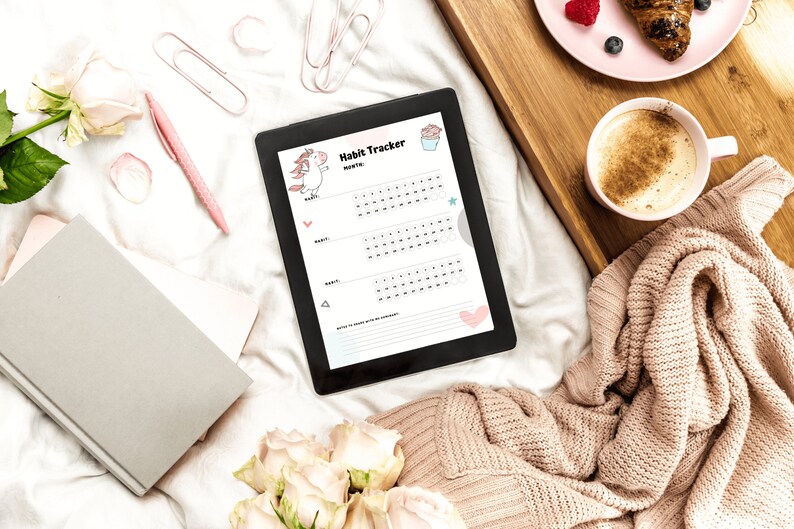 The digital habit tracker in the unicorn style of the monthly habit trackers for submissives - sitting on a bed next to coffee and a pink plate on a wooden tray, a knitted sweater, a gray notebook, and pink pen.