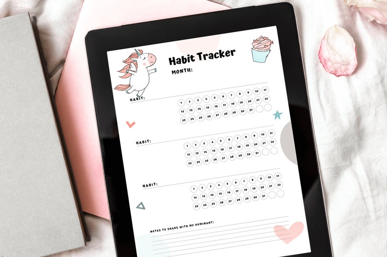 Digital version of the Unicorn Monthly Habit Tracker for submissives on a tablet next to gray and pink books and pink rose petals.