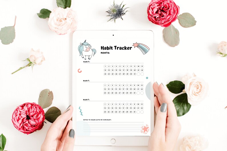 Digital habit tracker on an iPad - the unicorn monthly habit tracker for submissives. White hands with gray-green painted nails next to dark and light pink flowers and green leaves over white desk.