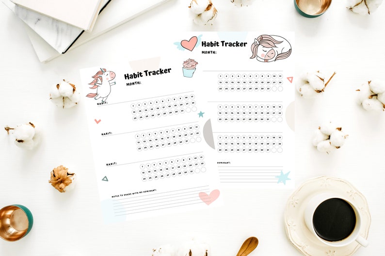 Two samples of the printed habit tracker in the Unicorn Monthly Habit Tracker sheets for submissives on a white desk next to a cup of coffee, flowers, and white notebooks.