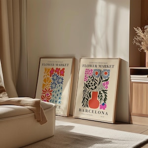 May include: Two framed art prints with floral designs. One features colorful flowers, the other a vase of flowers with the text "Barcelona". Both prints have a cream background and are in wooden frames. The prints are leaning against a wall.
