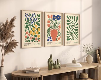 May include: Three framed art prints with floral designs in a Matisse style. The prints feature colorful flowers and foliage in vases and abstract patterns. The frames are a light wood color, and the prints are displayed on a white wall.