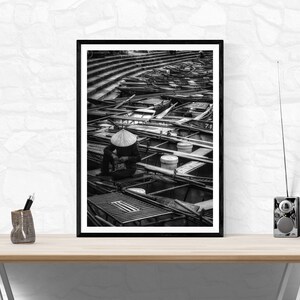 May include: Black and white framed photograph of a person wearing a conical hat, seated among numerous boats. The boats are arranged in a grid-like pattern, with a tiered structure in the background. The image is displayed on a wooden desk.