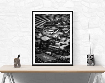 May include: Black and white framed photograph of a person wearing a conical hat, seated among numerous boats. The boats are arranged in a grid-like pattern, with a tiered structure in the background. The image is displayed on a wooden desk.