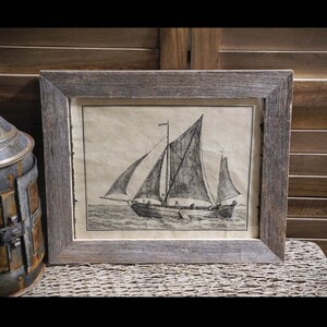 May include: A framed black and white print of a sailboat with a single mast and multiple sails. The print is set against a light brown background and is framed in a rustic wood frame.