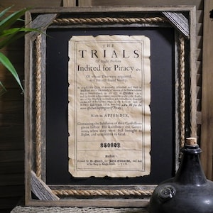 May include: A framed document with a black background and a white paper with the text "THE TRIALS Of Eight Perfons Indited for Piracy" and "With an APPENDIX, Containing the Substance of their Confessions given before His Excellency the Governor, when they were first brought to Goal." The document is framed with a rope border and a wooden frame.