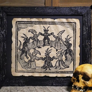 May include: A black framed print of a vintage illustration depicting a group of figures in a circle, possibly a dance or ritual. The figures are dressed in what appear to be medieval or renaissance-era clothing, with horns and other demonic features. The print is on a textured paper background.