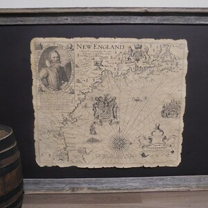 May include: Framed antique map of New England, featuring a portrait and intricate cartographic details. The map is printed on aged paper and set against a black background within a rustic, gray wooden frame. The map includes the text "New England".
