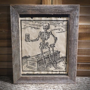 May include: A black and white woodcut print of a skeleton sitting on a coffin. The skeleton is holding an hourglass in its left hand and a scythe in its right hand. The print is framed in a rustic wooden frame.