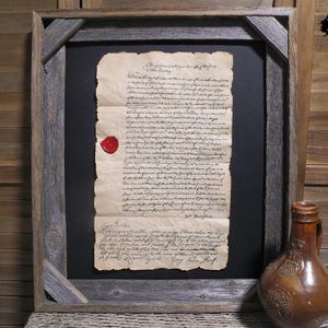 Op de afbeelding: Ingelijst antiek document met een rode waszegel. Het document is verouderd, met zichtbare slijtage. De lijst is gemaakt van rustiek, verweerd hout. Een bruine keramische kruik staat rechts van het frame.