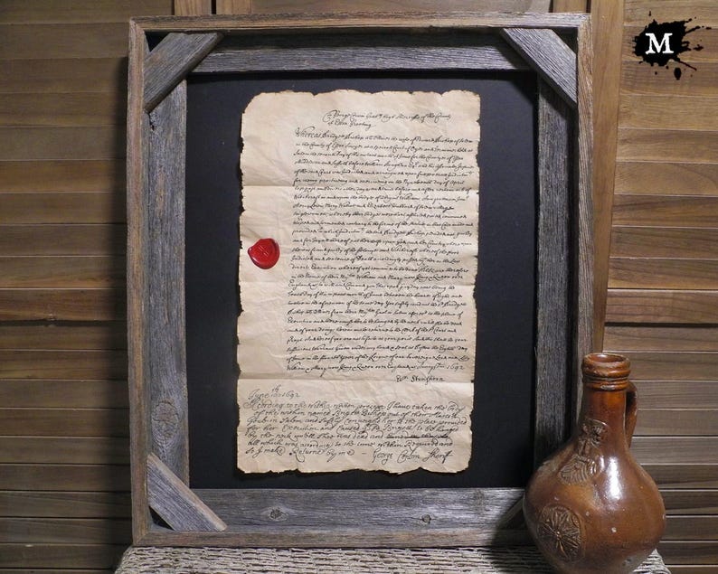 Op de afbeelding: Ingelijst antiek document met een rode waszegel. Het document is verouderd, met zichtbare slijtage. De lijst is gemaakt van rustiek, verweerd hout. Een bruine keramische kruik staat rechts van het frame.