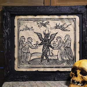 May include: A black and white woodcut print depicting a devil with wings and a tail, holding a small doll. Four people, two women and two men, are standing around the devil, looking at the doll. The print is framed in a black frame.
