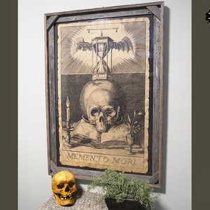 May include: A framed black and white print of a skull with an hourglass above it. The print is on a textured paper and has the words "Memento Mori" at the bottom. The frame is made of rustic wood.