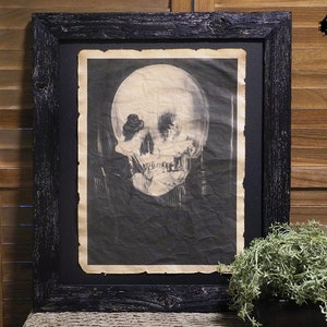 May include: A black framed print of a skull with a textured background. The print is in a black frame with a distressed finish.