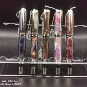 May include: A collection of five fountain pens displayed on a clear acrylic stand. The pens feature silver accents and colorful, marbled barrels in shades of blue, orange, brown, pink, and burgundy. Each pen has a silver clip and a nib.