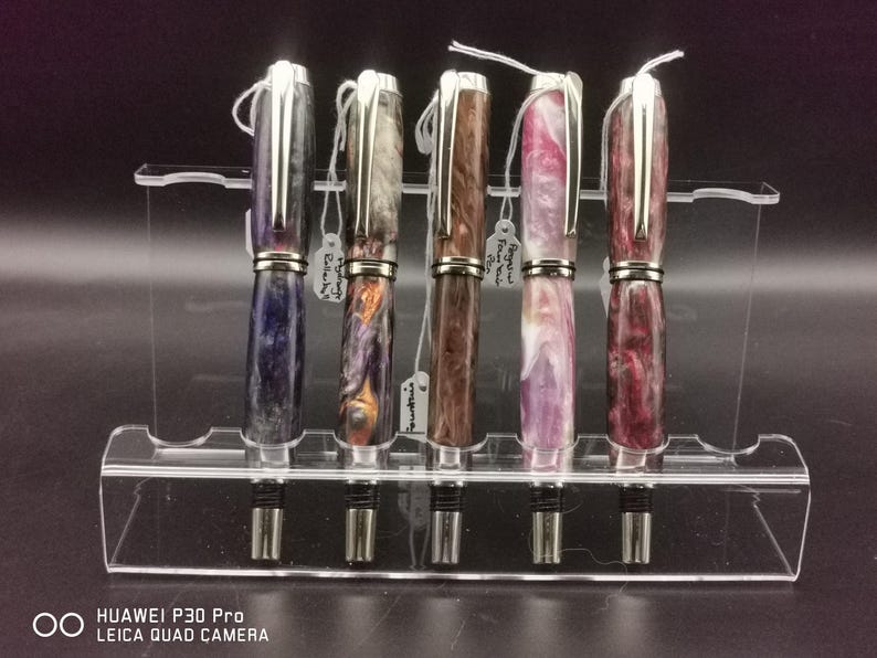 May include: A collection of five fountain pens displayed on a clear acrylic stand. The pens feature silver accents and colorful, marbled barrels in shades of blue, orange, brown, pink, and burgundy. Each pen has a silver clip and a nib.