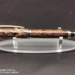 May include: A handcrafted pen with a marbled brown and cream barrel, silver accents, and a silver clip. The pen is displayed on a clear acrylic stand, showcasing its elegant design. The pen's tip is silver.
