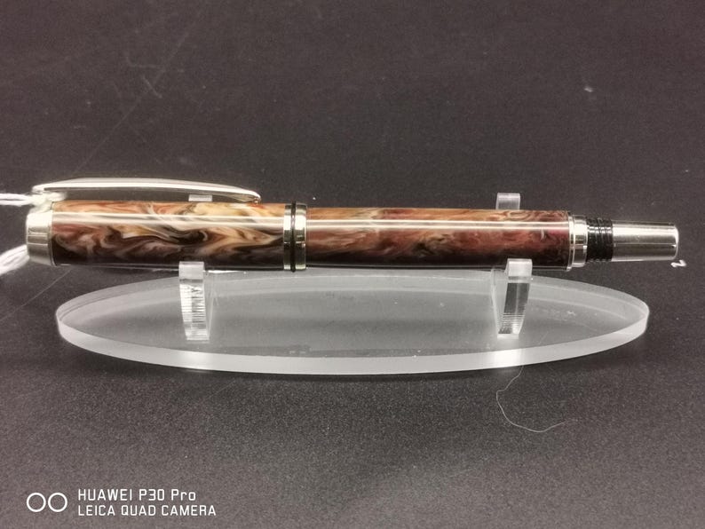 May include: A handcrafted pen with a marbled brown and cream barrel, silver accents, and a silver clip. The pen is displayed on a clear acrylic stand, showcasing its elegant design. The pen's tip is silver.