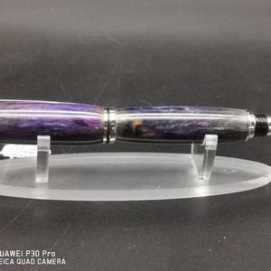 May include: A handcrafted pen with a purple and gray marbled design. The pen has a silver clip and tip, and is displayed on a clear acrylic stand. The pen's body features a swirling pattern of colors.