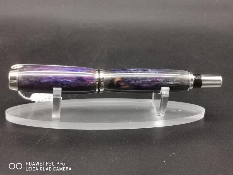 May include: A handcrafted pen with a purple and gray marbled design. The pen has a silver clip and tip, and is displayed on a clear acrylic stand. The pen's body features a swirling pattern of colors.