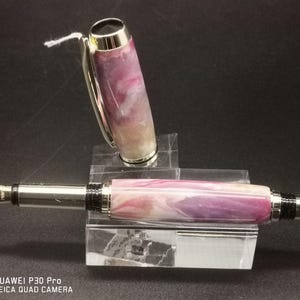May include: A fountain pen with a marbled purple, pink, and white barrel and silver accents. The cap is off, revealing the nib. The pen rests on a clear acrylic stand. A small white tag is attached to the cap.