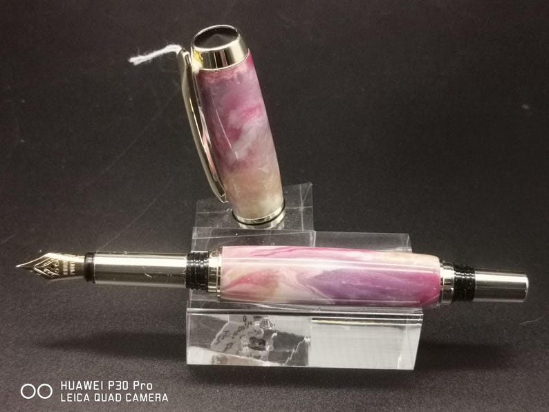 May include: A fountain pen with a marbled purple, pink, and white barrel and silver accents. The cap is off, revealing the nib. The pen rests on a clear acrylic stand. A small white tag is attached to the cap.