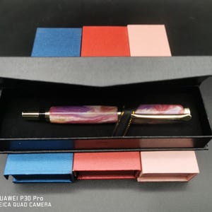 May include: A luxury fountain pen with a marbled purple and gold barrel, presented in a black box. The pen features a gold-colored clip and nib. The box is set against a backdrop of small blue, red, and pink boxes.