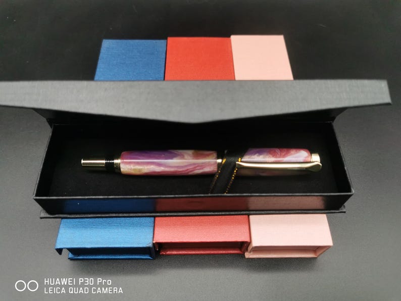 May include: A luxury fountain pen with a marbled purple and gold barrel, presented in a black box. The pen features a gold-colored clip and nib. The box is set against a backdrop of small blue, red, and pink boxes.