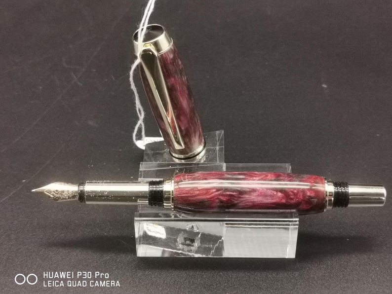May include: A fountain pen with a burgundy and white marbled barrel and silver accents. The cap is off, revealing the nib. The pen is displayed on a clear acrylic stand. The pen is a writing instrument.