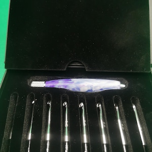 May include: A set of crochet hooks presented in a black box. The hooks are silver with green detailing and size markings. A larger hook with a purple and white marbled handle is also included. The box is lined with black velvet.