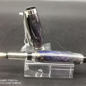 May include: A fountain pen with a silver nib and accents. The pen's body features a swirling pattern of purple, blue, and gray. The cap is open, revealing the nib. The pen is displayed on a clear acrylic stand.
